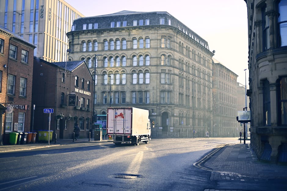 A white Iveco moving van positioned on a narrow urban street between residential buildings with historic facades, some with balconies and decorative wrought iron railings. The van is backed up close t