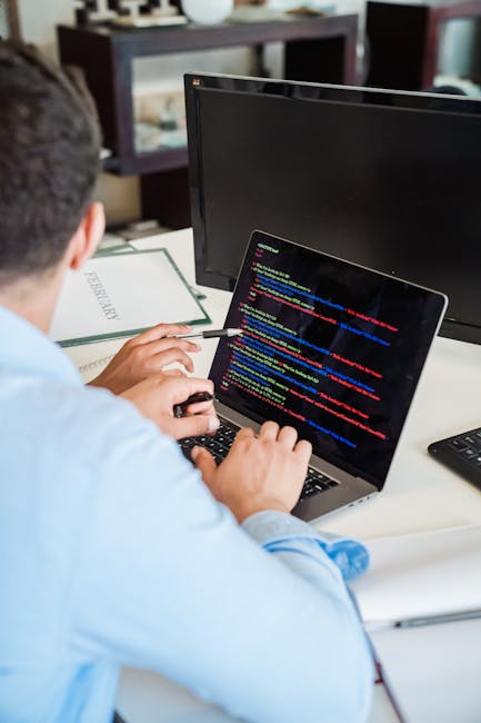 A close-up view of a black laptop computer with a glowing red-backlit keyboard, displaying lines of code on the screen, placed on a dark surface near a white Puma-branded baseball cap. In the backgrou